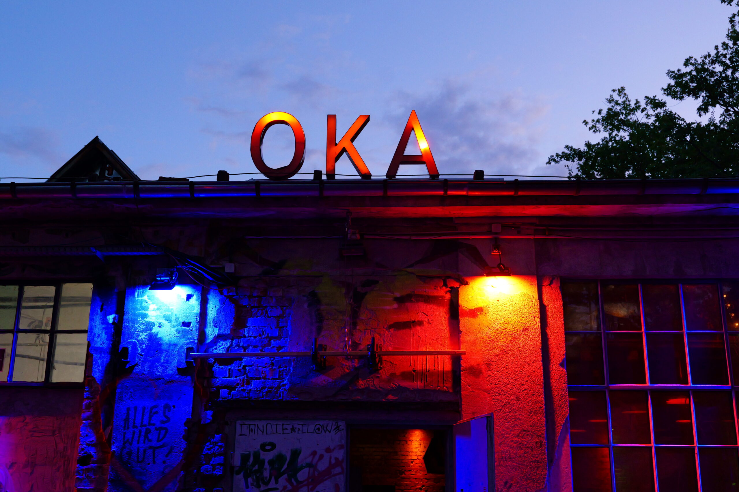 Industriegebäude mit dem leuchtenden Schriftzug „OKA“, beleuchtet in Blau- und Rottönen bei Abenddämmerung.
